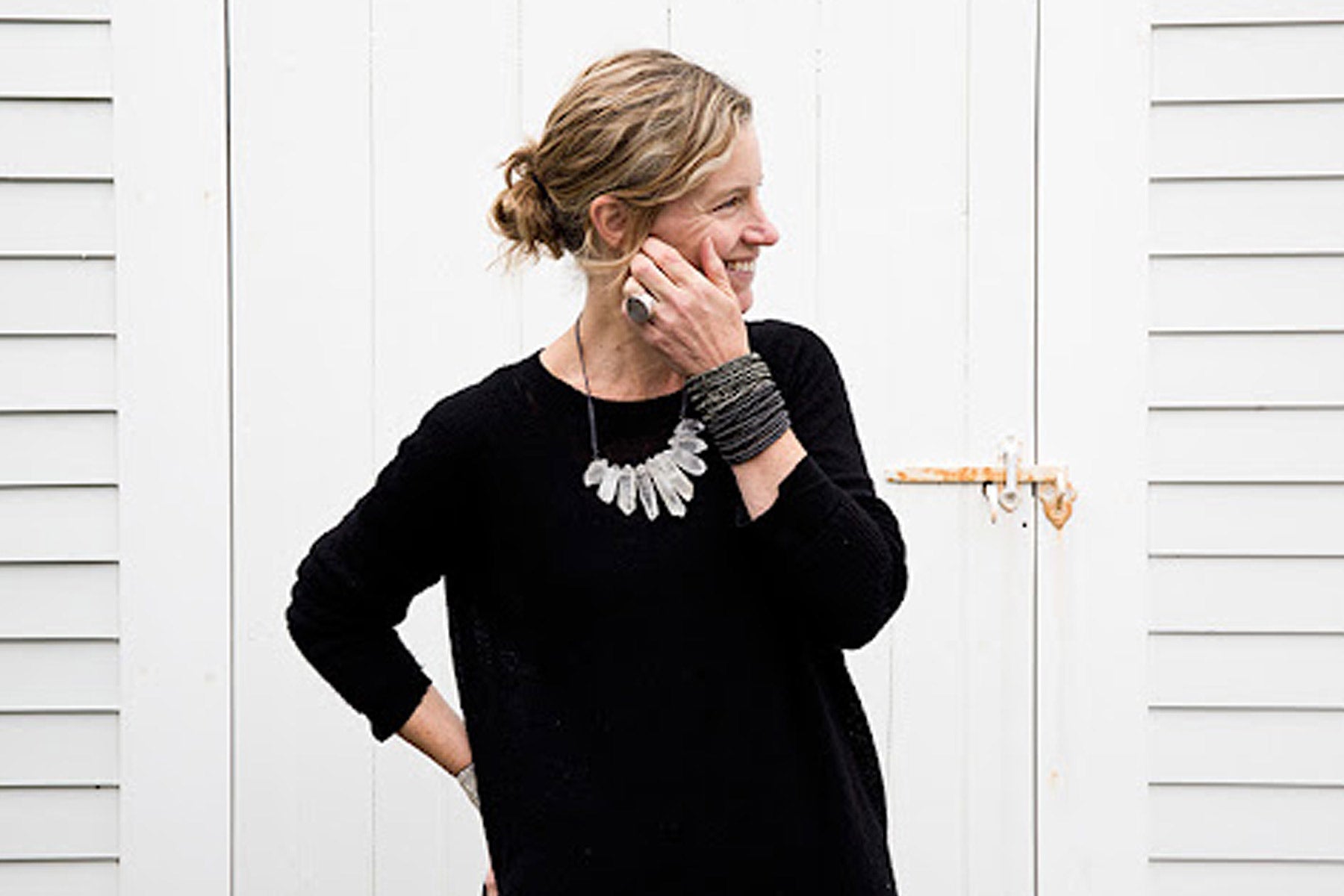 A woman with light hair in a loose bun stands sideways, smiling and touching her face. She wears a black sweater, chunky bracelets, and a statement necklace, standing in front of white wooden doors with a rusty latch.