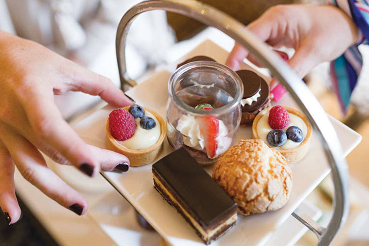 Two hands reaching for assorted desserts on a tiered tray, including fruit-topped tarts, a cream-filled pastry, chocolate-layered cake, and a jar with whipped cream and strawberries.