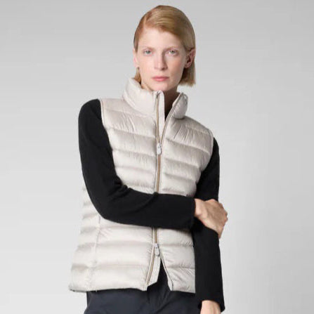 A woman with long brown hair is wearing a white sweater, black shorts, white socks, and black loafers. She is also sporting oversized square glasses and the SAVE THE DUCK - LYNN PUFFER VEST from SAVE THE DUCK made from quilted technical fabric. She stands against a plain gray background.