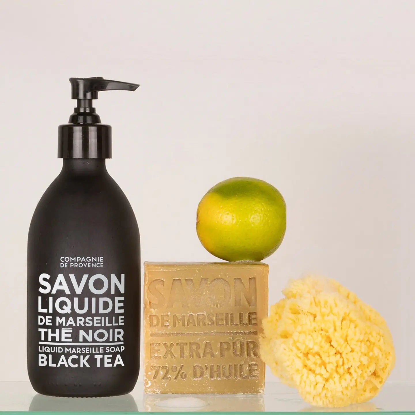 A black pump bottle of liquid Marseille soap, a bar of solid Marseille soap, a yellow sea sponge, and a lime are arranged on a light surface against a plain background.