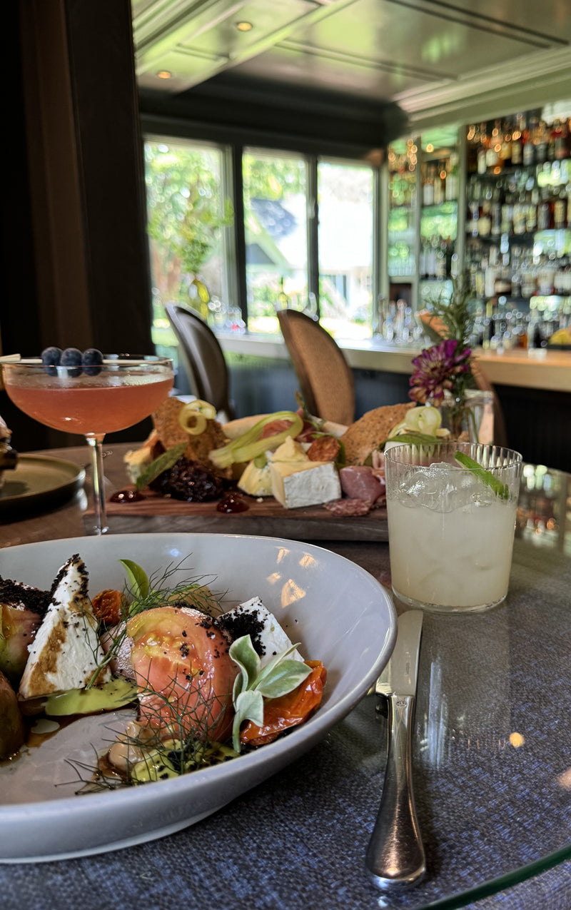 A table set with a cocktail, a glass of lemonade, a goat cheese and tomato salad, and a charcuterie board, with a stylish bar and large windows in the background.
