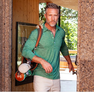 A man with short gray hair and a beard, wearing the CRIQUET - BRRR° Long Sleeve Range Polo in Reed and light pants, stands in a doorway holding a strap with golf clubs and accessories over his shoulder.
