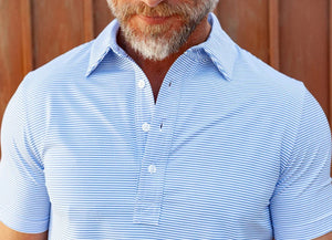 A man wears the CRIQUET Performance Sport Range Polo in Hobby Stripe - Bluebonnet, featuring light blue and white stripes, a collared neckline with three buttons, and lightweight, breathable fabric, standing against a plain white background.