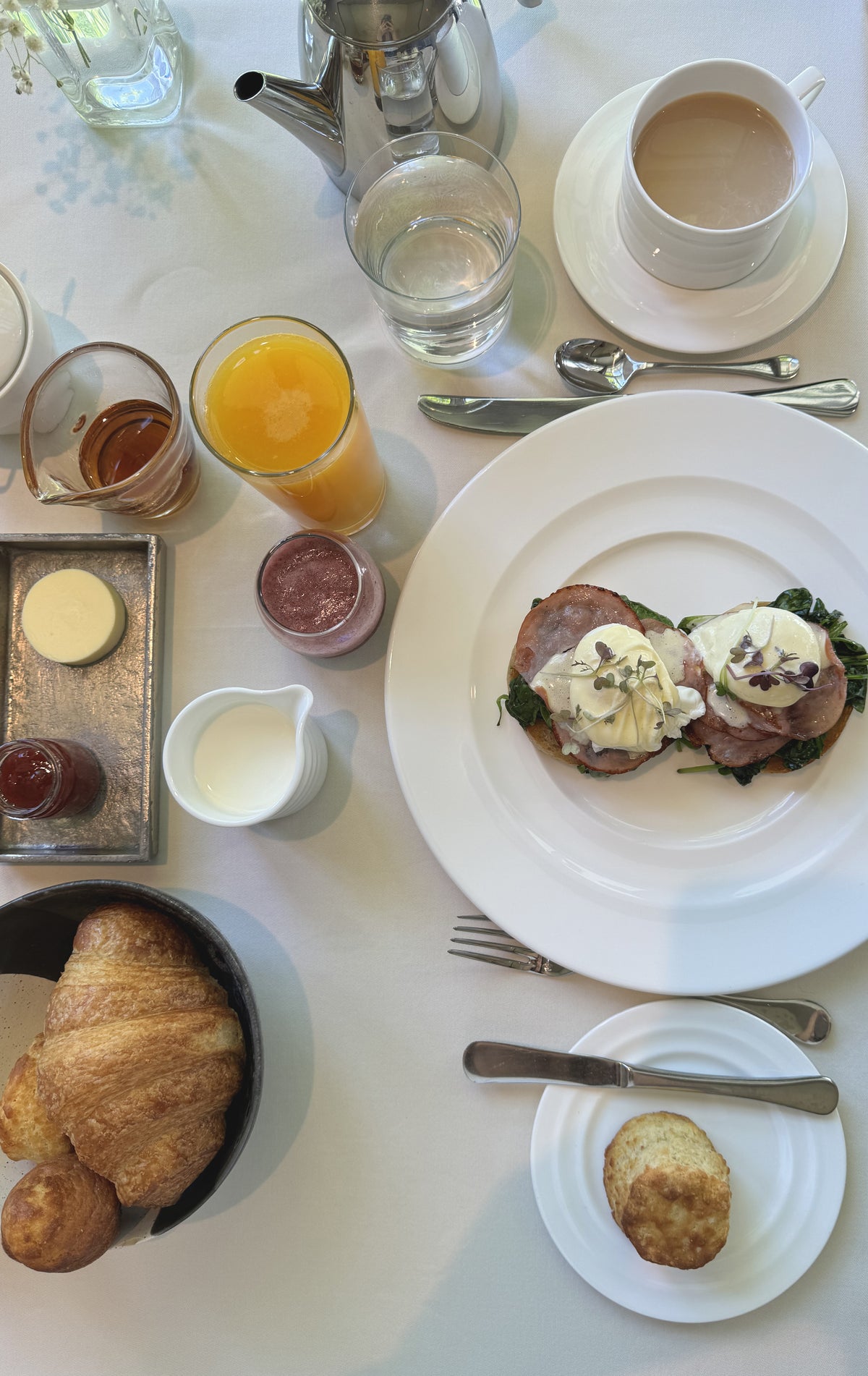 A breakfast table set with eggs Benedict on a plate, a cup of coffee, orange juice, water, croissants in a bowl, a scone on a small plate, butter, jam, and cream, all arranged neatly on a white tablecloth.