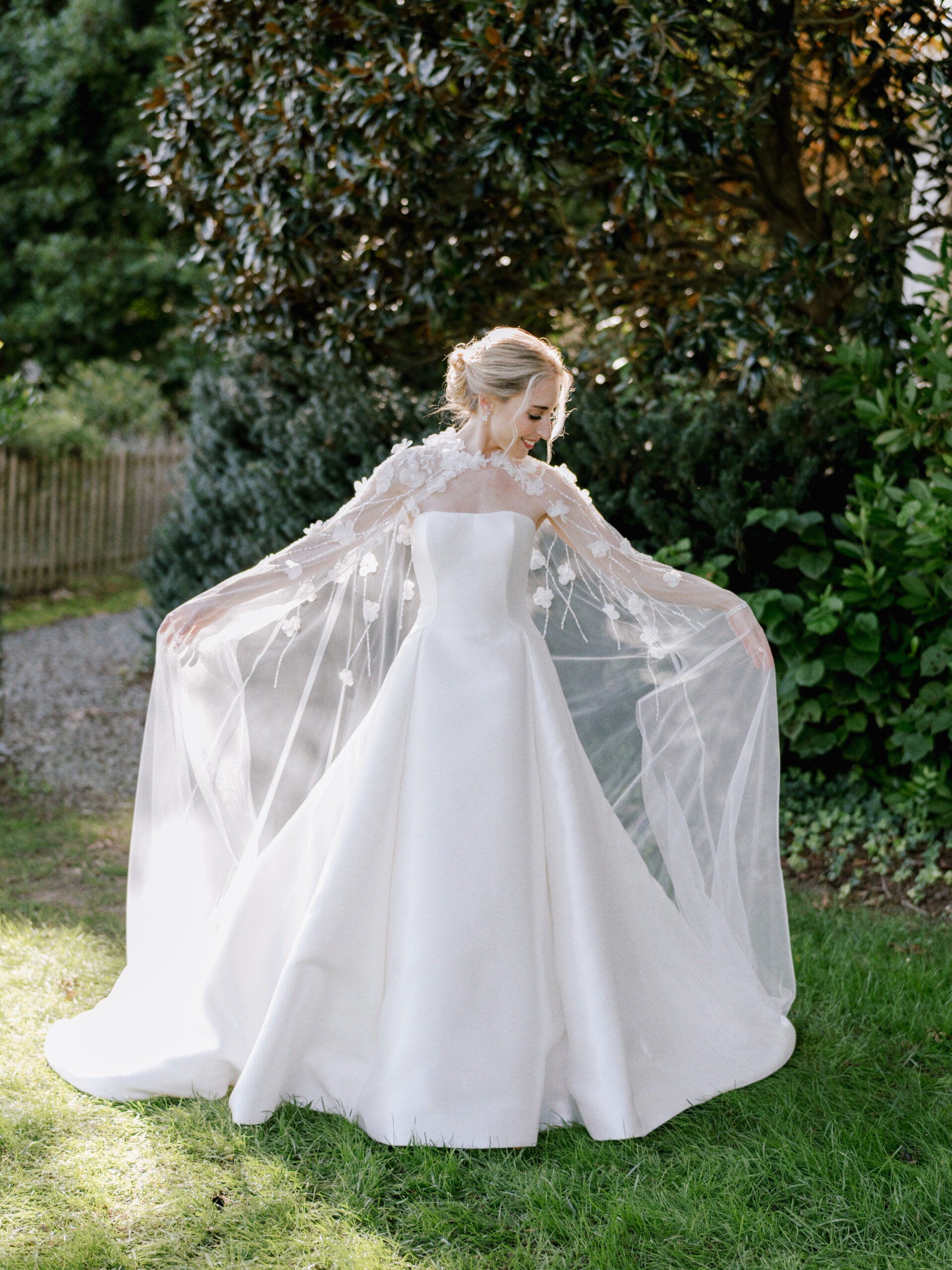 A bride stands outdoors in sunlight, wearing a white gown with a flowing sheer cape decorated with floral appliqués. She holds the cape out, creating a wing-like effect, with greenery and trees in the background.