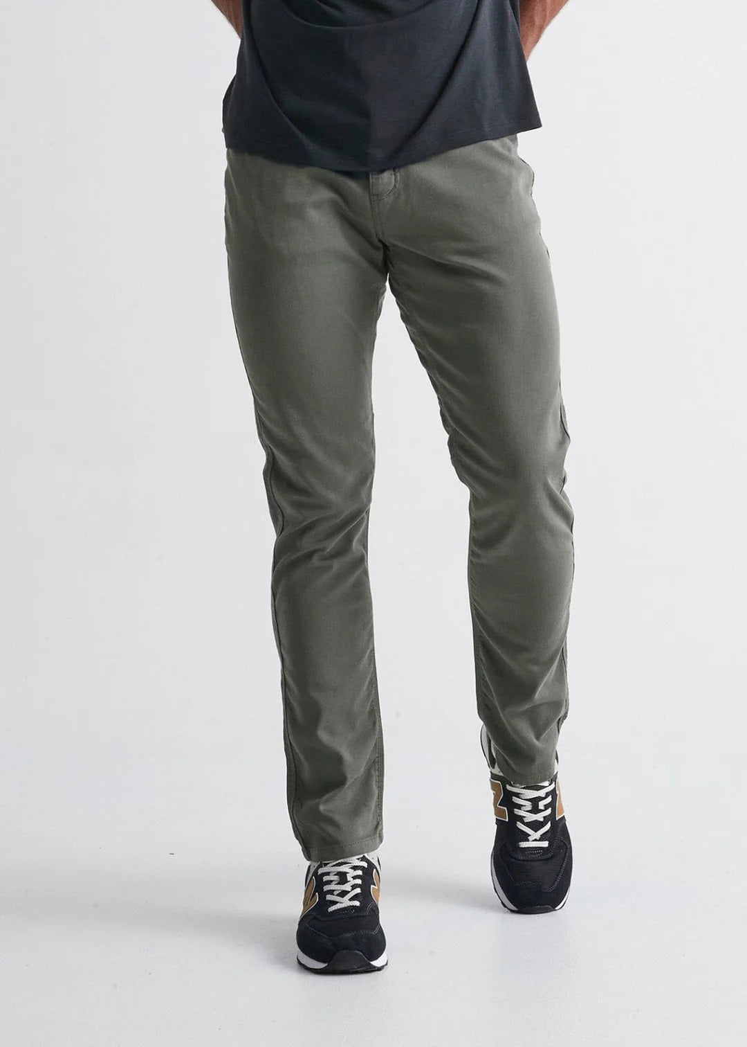 A person wearing olive-green pants, a black t-shirt, and black sneakers with white laces stands against a plain white background. The person's head and upper torso are not visible.