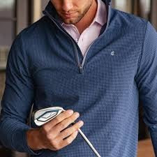 A man in the CRIQUET Seersucker Performance Pullover in Navy and light pants smiles with his hand by his side; this lightweight layer from CRIQUET offers UPF 50 protection against a simple background.