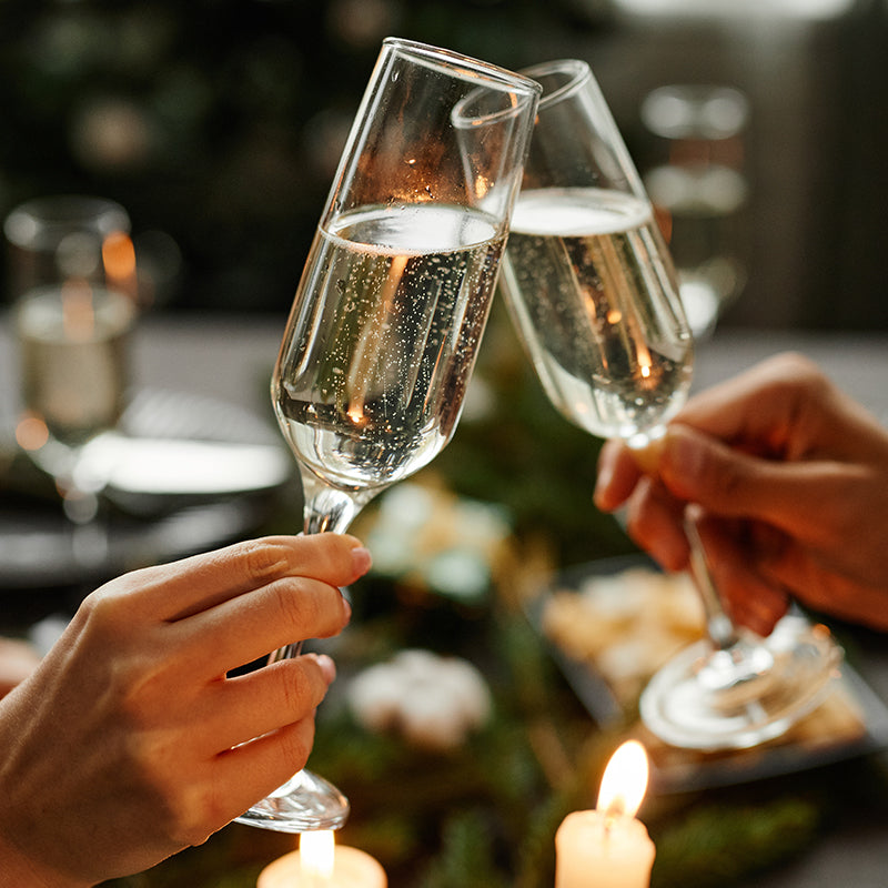 Two people clink champagne glasses in a toast over a candlelit table, with blurred food and drinks in the background, creating a festive and celebratory atmosphere.