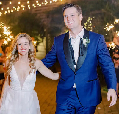 A bride in a white dress and a groom in a blue suit walk hand in hand, smiling, under string lights at night, surrounded by guests holding sparklers.