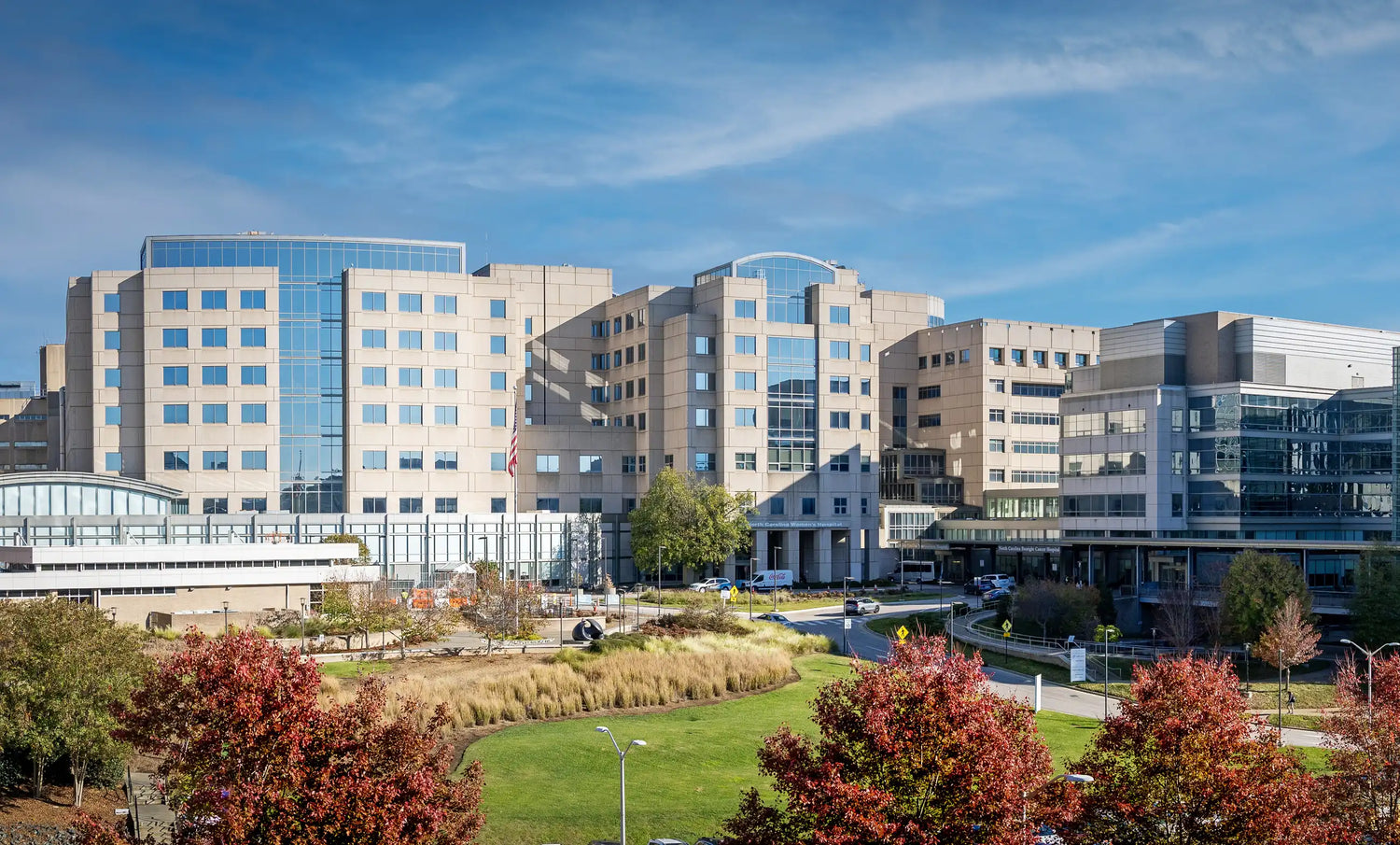 A large modern hospital complex with multiple beige buildings, large windows, and glass walkways, surrounded by landscaped green lawns and trees with autumn foliage under a blue sky.