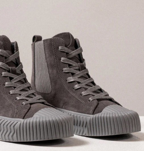 A pair of DANIELLA LEHAVI - OSAKA HIGH TOP SNEAKERS made from gray suede with matching gray rubber soles and toe caps, displayed on a white platform against a neutral background. These comfortable sneakers from DANIELLA LEHAVI feature a lace-up design and ribbed detailing on the sides and soles, showcasing their contemporary design.
