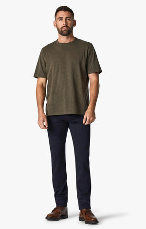 A man with short hair and a beard stands facing forward, wearing an olive green T-shirt, 34 HERITAGE - COURAGE STRAIGHT LEG PANT IN NAVY TWILL, and brown shoes against a plain white background.