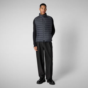 A man wearing the SAVE THE DUCK - RHUS VEST with Recycled Plumtech padding over an olive cardigan, paired with loose-fitting olive pants and brown shoes, stands against a plain light background with his hand in his pocket.