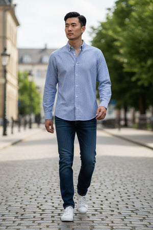 A man in a FRANK & EILEEN Finbar Men's Button Up Shirt in Blue Stripes, blue jeans, and white sneakers walks confidently down a sunny, tree-lined cobblestone street.