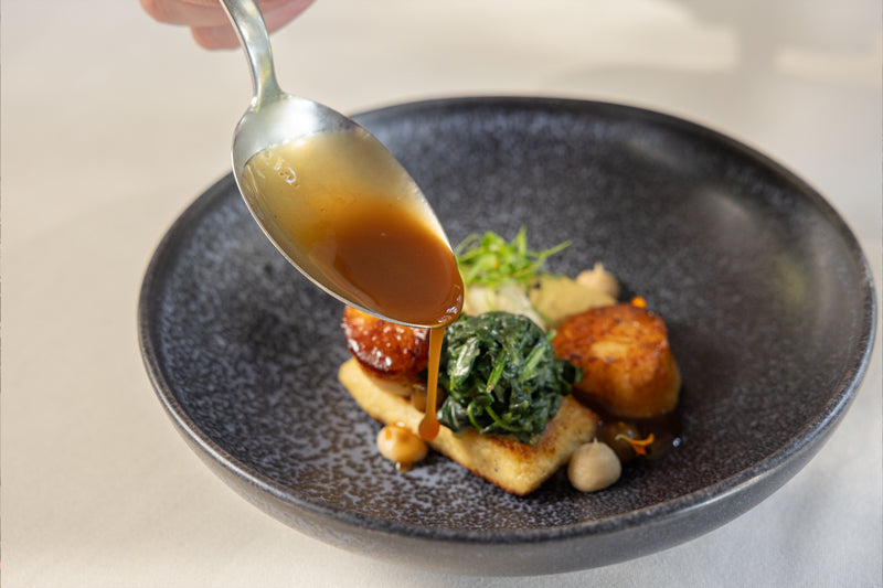 A hand holds a spoon, drizzling brown sauce over a gourmet dish with seared scallops, sautéed greens, chickpeas, and a golden-brown base, all served on a dark, textured plate.