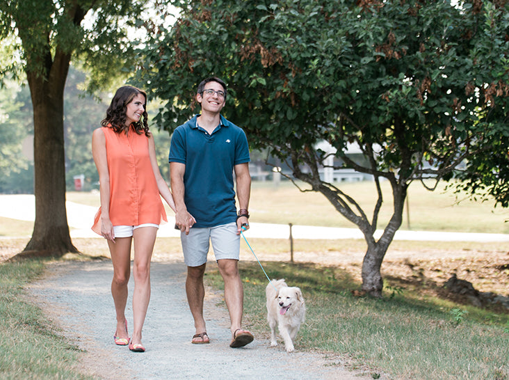 A smiling couple walks a fluffy white dog on a leash along a tree-lined path in a park. The woman wears a sleeveless orange top and shorts, and the man is in a blue polo and shorts. It’s a sunny day.