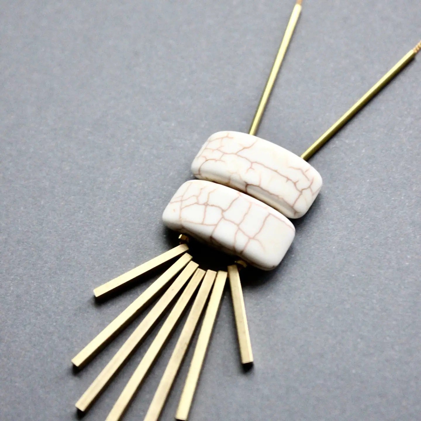 A necklace with two white, crackled oval beads stacked together, accented by several slim, gold metal bars fanning out below on a gray background.