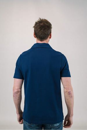 A man with light brown hair and a short beard wears the EDWARD ARMAH Pima Cotton Tailored Polo Shirt in dark blue and jeans, standing against a plain light background with a relaxed expression and one hand in his pocket.