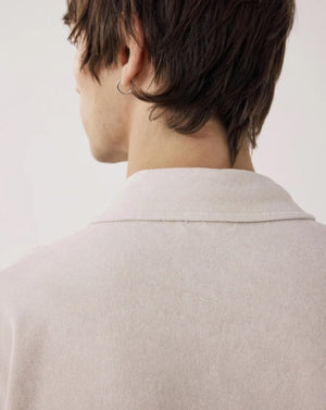 Wearing the HARTFORD Terry Polo Shirt in Stone with sage green cargo pants, a person stands with one hand in their pocket, glancing to the side against a plain background.