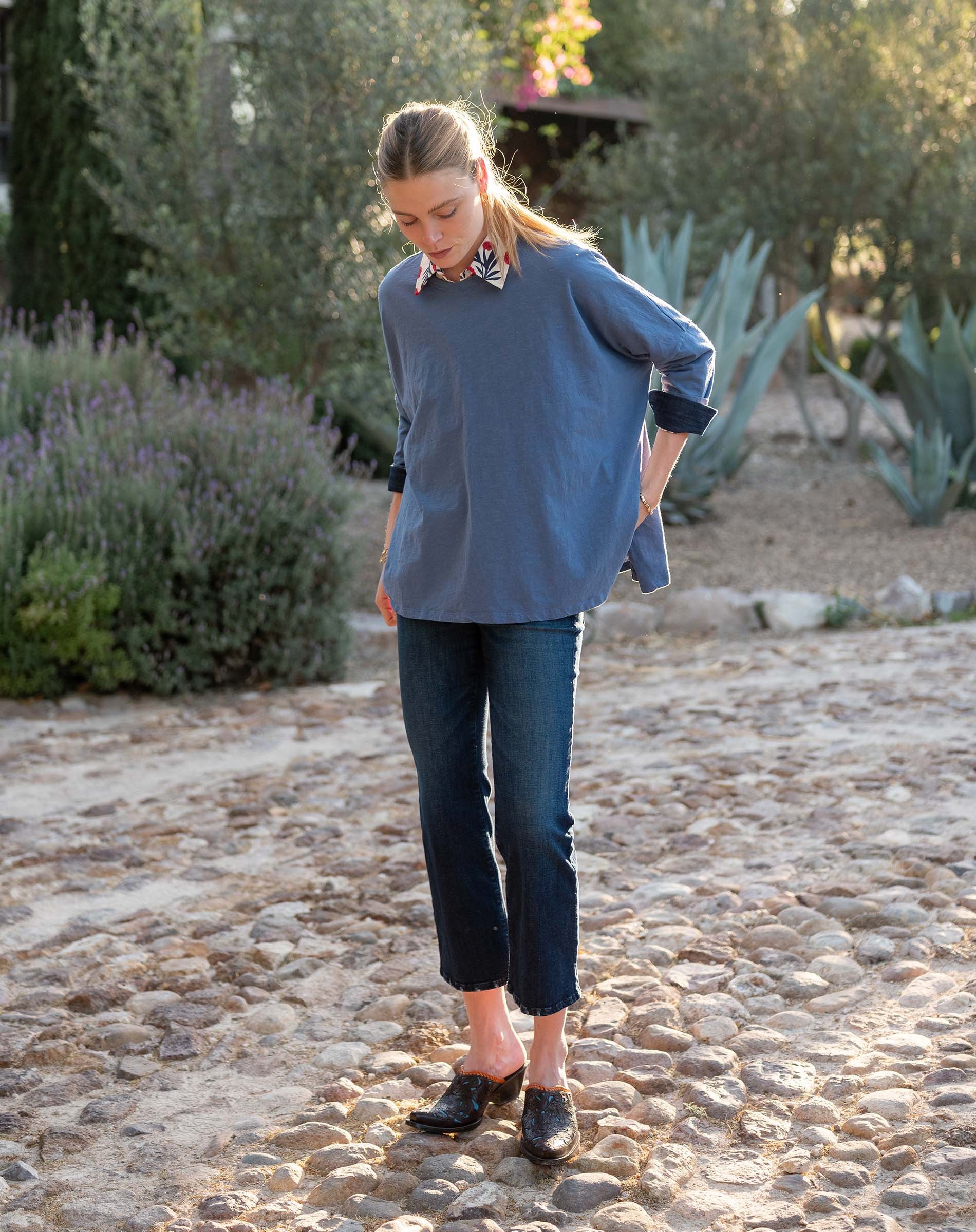 A woman stands outdoors in the MERSEA - CATALINA SLUB TEE IN MOON BLUE by MERSEA, a floral-patterned collar peeking out as she smiles softly amid sunlight and greenery.