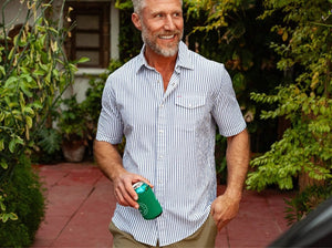 A smiling man with gray hair and a beard wears a short-sleeved, white and blue striped shirt and khaki pants. He stands outdoors holding a green mug, with greenery and a red-tiled patio in the background.