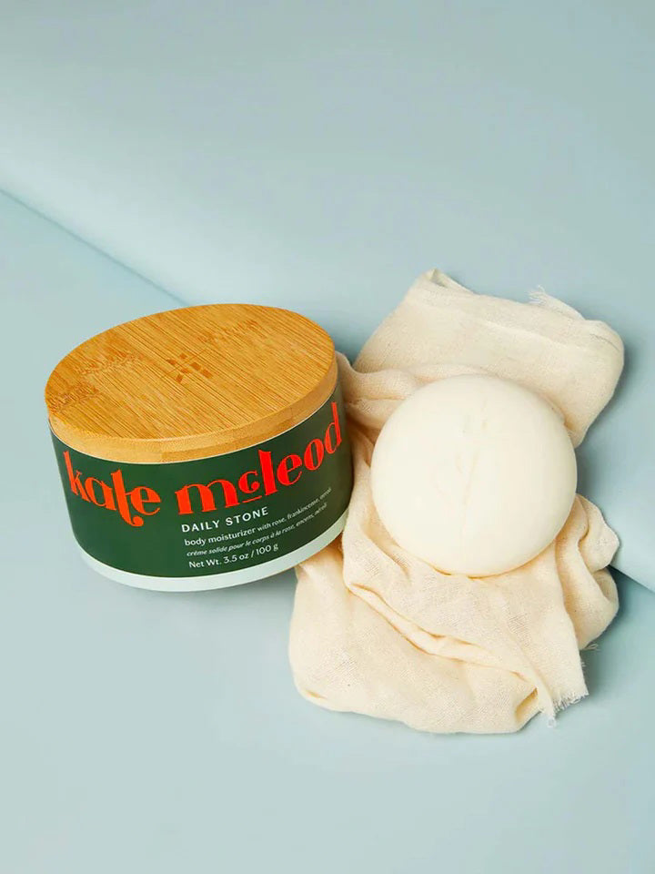 A KATE MCLEOD - DAILY STONE DEEP MOISTURIZING LOTION BAR with a wooden lid rests beside a luxurious Body Stone on beige cloth against a blue backdrop, promising daily indulgence.