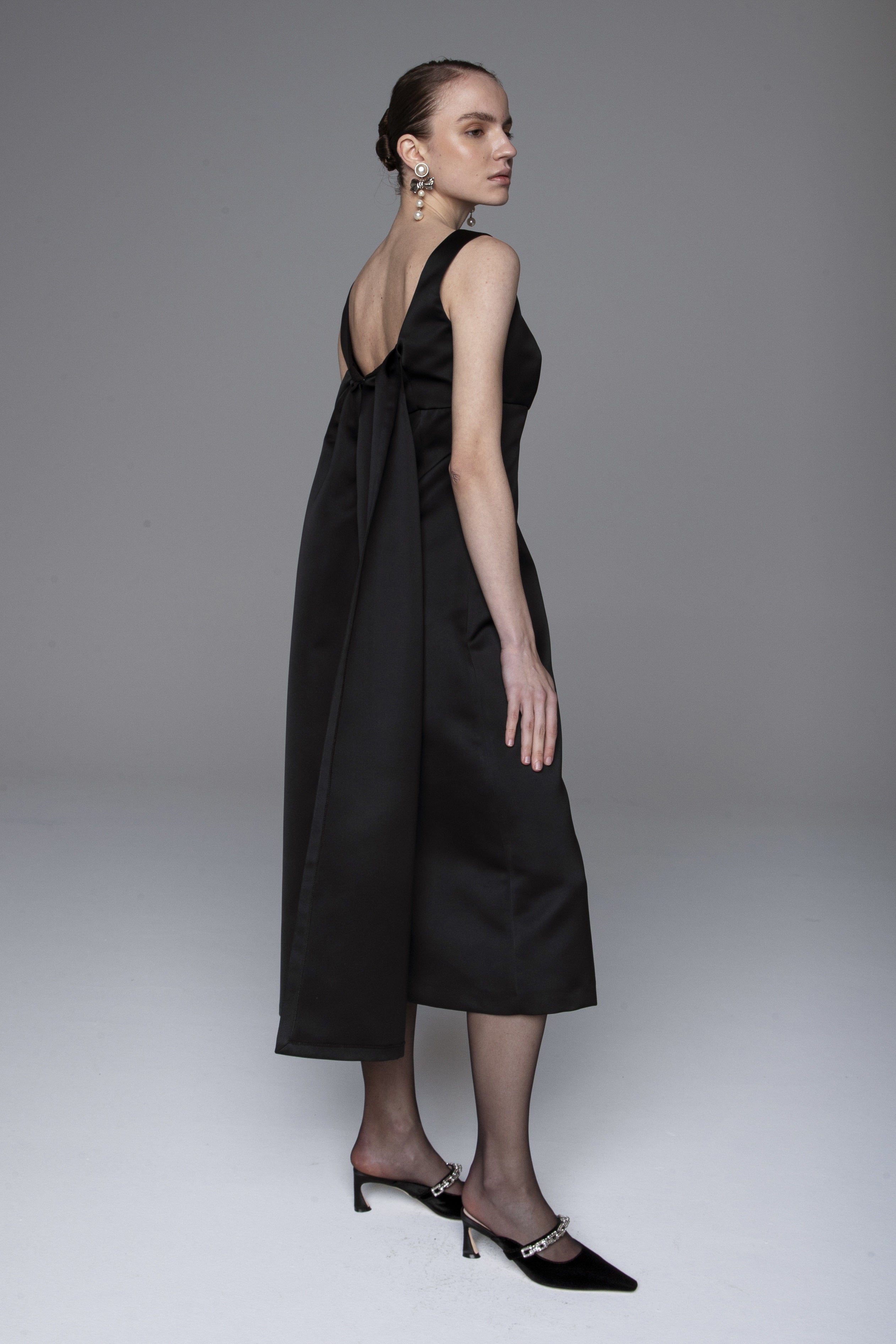 A woman stands posing in a sleeveless, black, mid-length dress with a deep V-back and large bow detail. She wears black pointed heels with embellishments and statement earrings, with her hair pulled back. The background is plain gray.