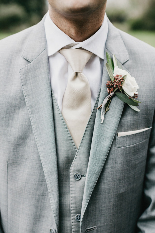 A man dressed in a light gray suit, white shirt, and beige tie, with a white rose boutonniere pinned to his lapel. The photo is focused on his chest and torso, not showing his full face.