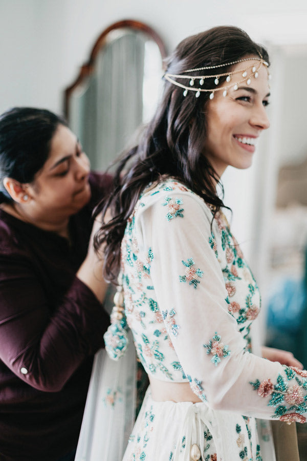 A smiling woman in a white floral embroidered outfit and pearl headpiece is getting her hair styled by another woman. They are indoors, standing near a mirror.