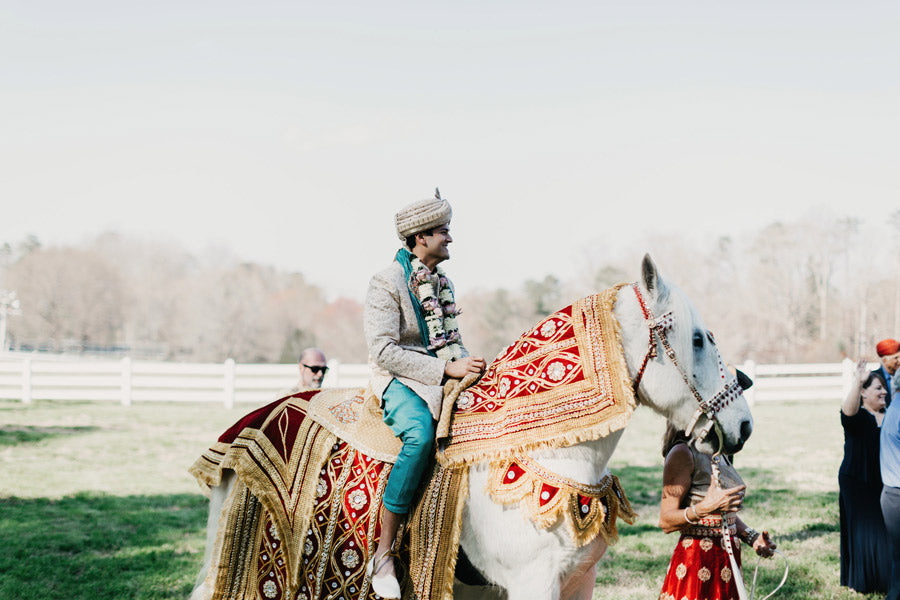 A groom in traditional Indian attire and a turban sits on a white horse draped in ornate red and gold fabric during an outdoor wedding ceremony, with people and trees in the background.