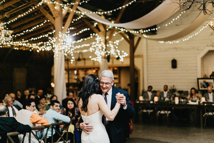 A bride in a white dress dances with an older man in a suit at a warmly lit wedding reception. Guests watch from tables, and string lights hang from the rustic wooden ceiling.