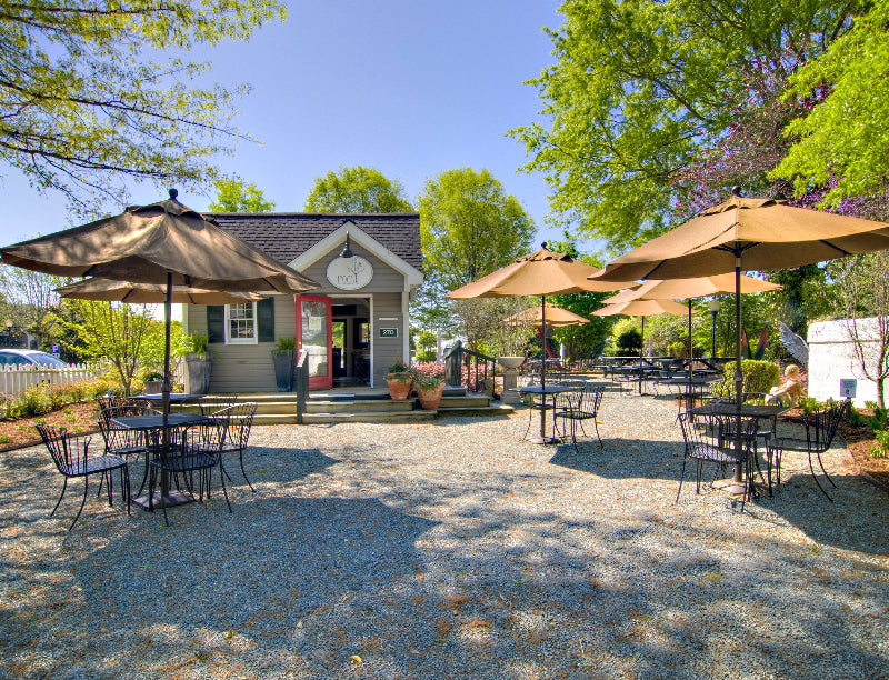 Outdoor cafe with black metal tables and chairs, large tan umbrellas, and a small gray building with a pink door in the background, surrounded by green trees and plants on a sunny day.