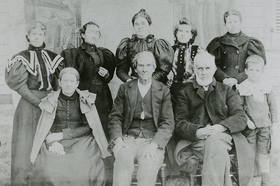 A black-and-white vintage photo of eight adults and one boy, dressed in late 19th-century clothing, posing together in two rows: five women standing, three seated adults, and a boy standing on the far right.