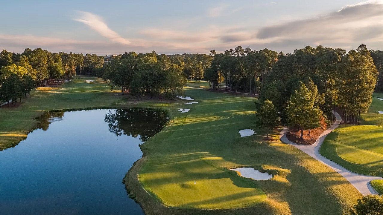 Aerial view of a scenic golf course with lush green fairways, sand bunkers, a reflective pond, and trees lining the landscape under a partly cloudy sky at sunset.