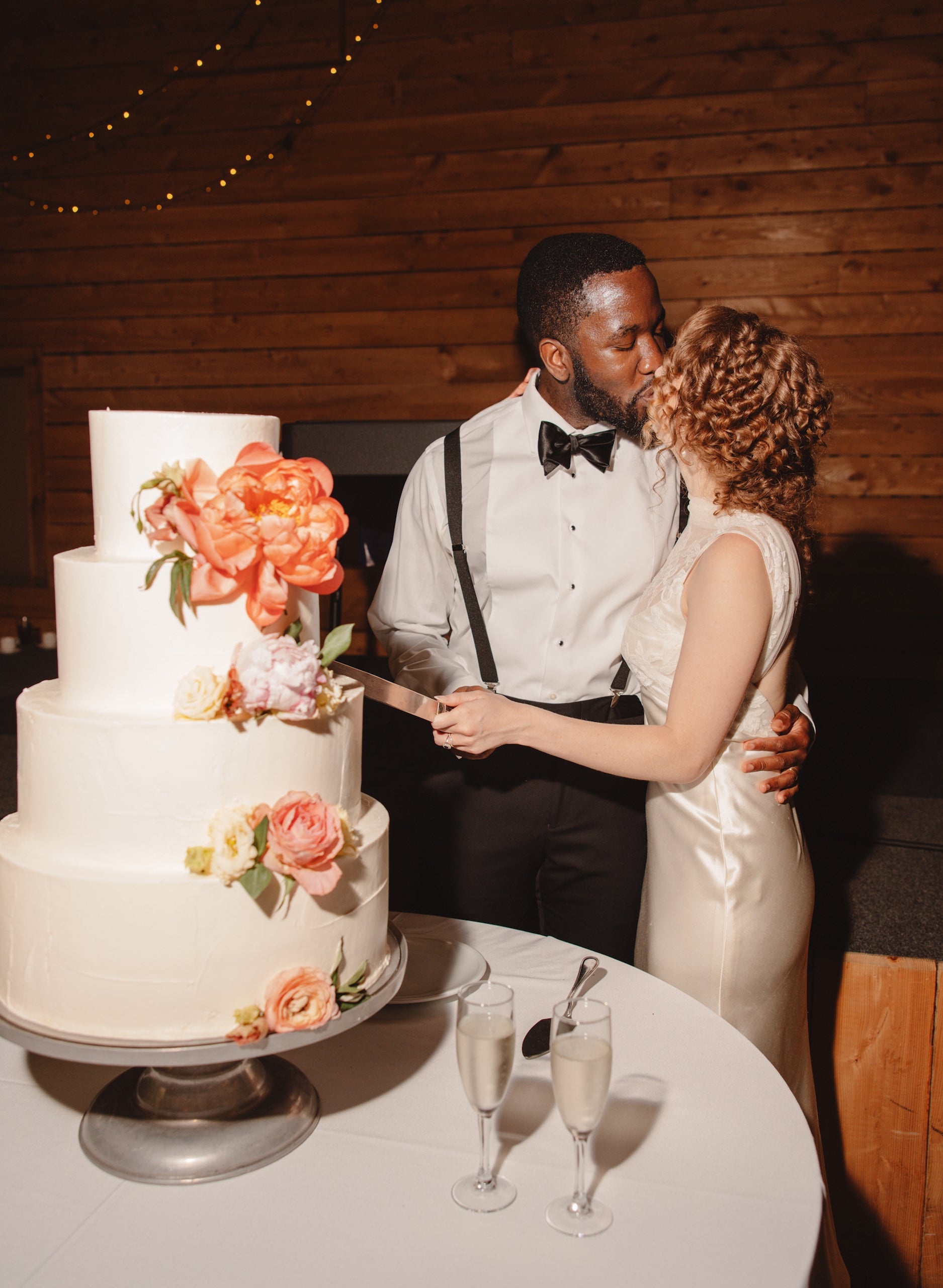 A couple dressed in formal wedding attire shares a kiss while cutting a three-tiered white wedding cake decorated with colorful flowers. Two glasses of champagne sit on the table beside the cake.
