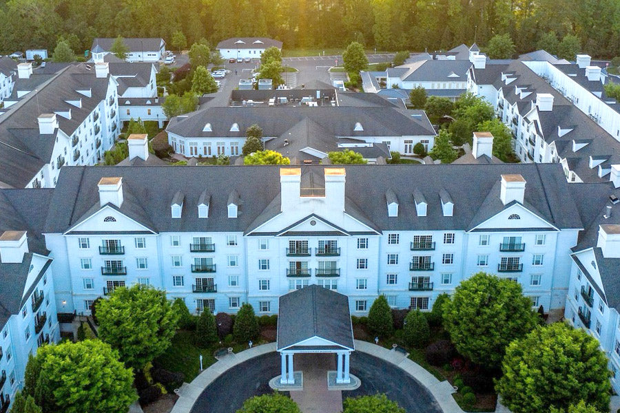 Aerial view of a large, elegant white building complex with dark roofs, surrounded by green trees and landscaped grounds, taken at sunset with sunlight filtering through the background trees.