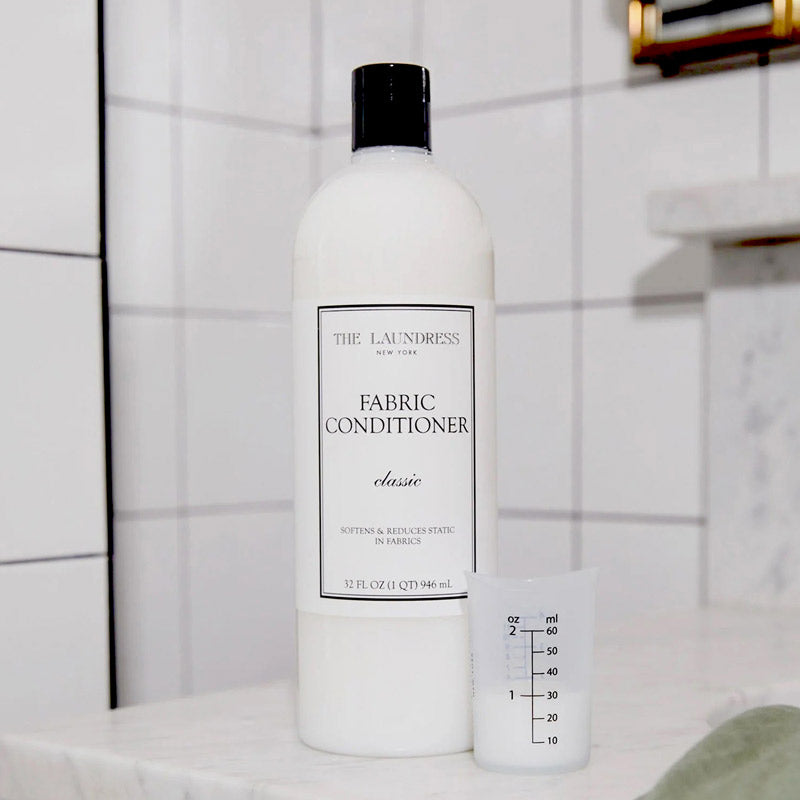 A bottle of The Laundress New York Fabric Conditioner stands on a white marble surface, with a clear measuring cup beside it. The background features white tile and part of a faucet.