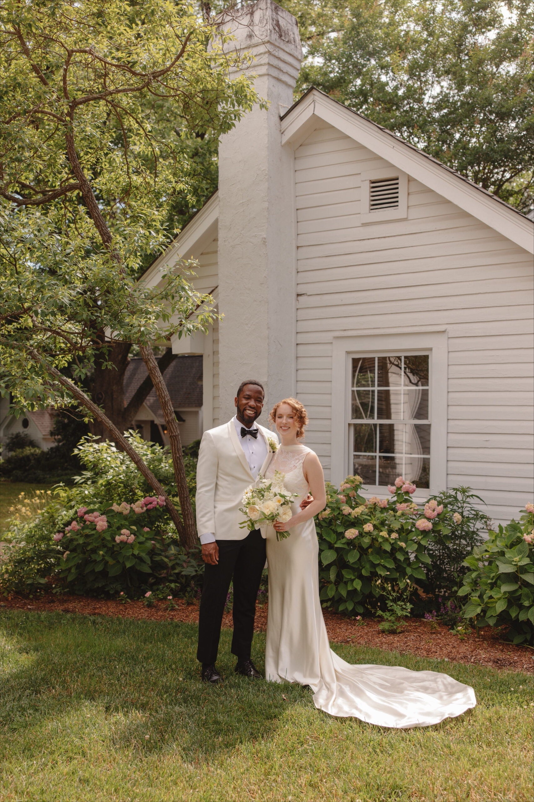 A newlywed couple stands smiling in front of a white house with flowering bushes. The groom wears a white tuxedo jacket and black pants; the bride wears a sleeveless white gown and holds a bouquet.