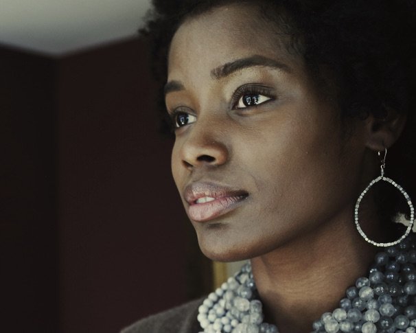 A woman with short curly hair gazes thoughtfully out of frame. She wears a beaded necklace and large hoop earrings, with soft natural light illuminating her face.