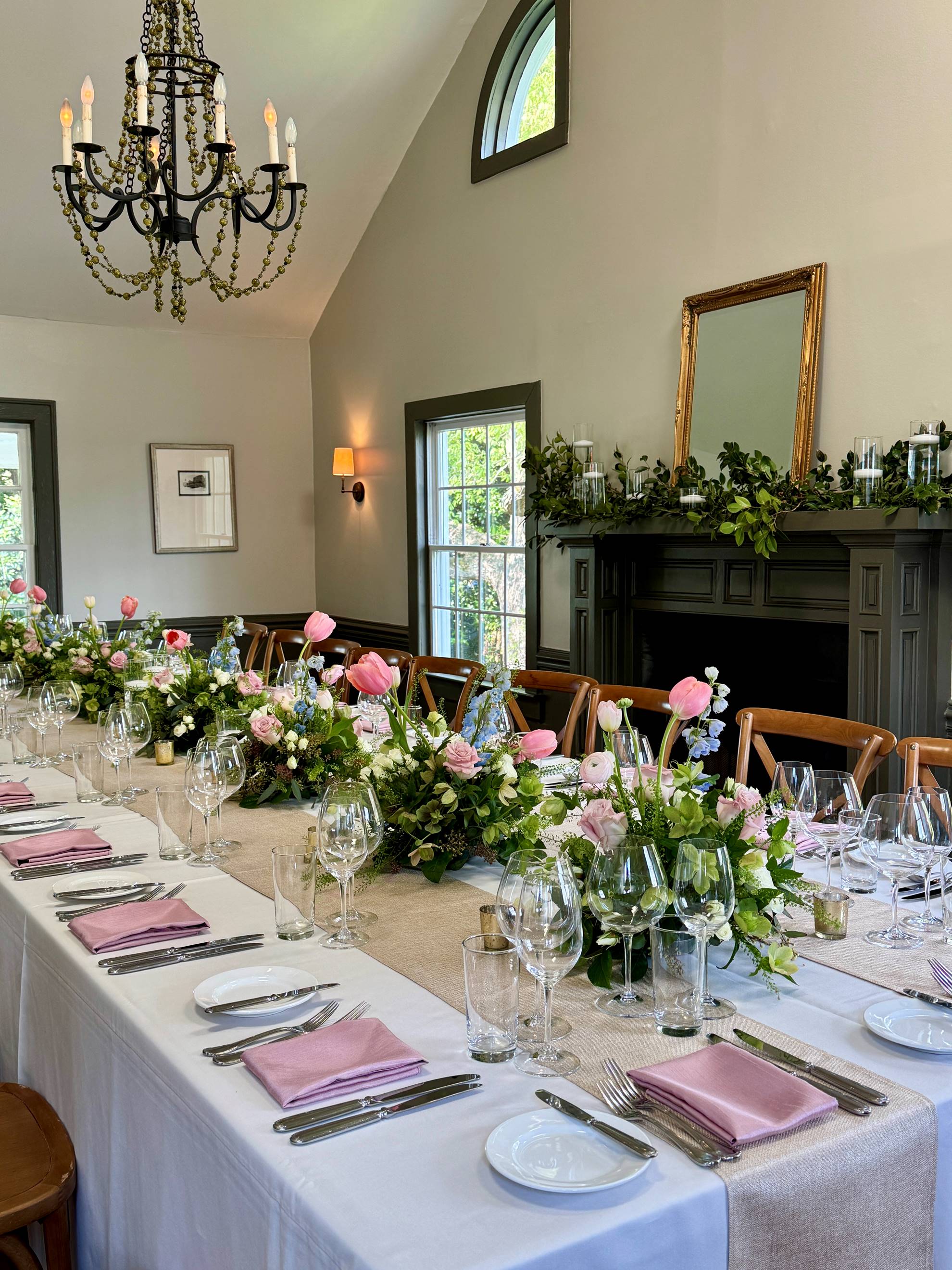 Elegant dining table set for a formal event, decorated with pink and white floral centerpieces, pink napkins, glassware, and cutlery. Room features a chandelier, greenery on a mantle, and large windows.