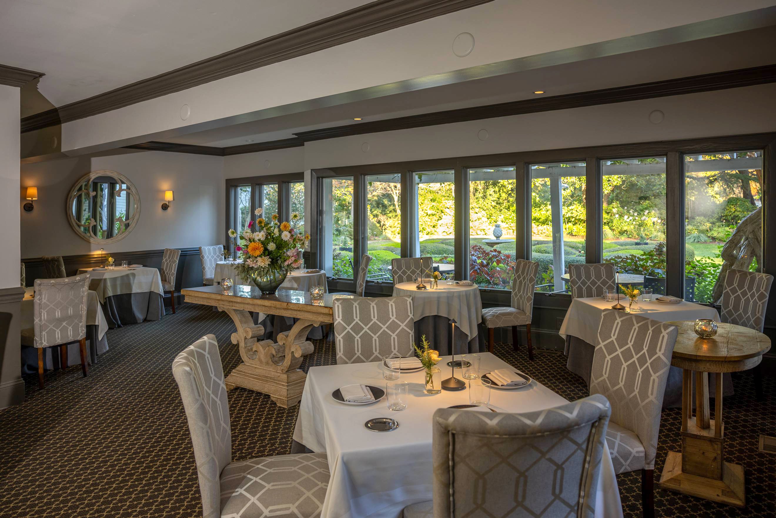 Elegant restaurant dining room with white tablecloths, patterned chairs, large windows overlooking a lush garden, soft lighting, and a central table decorated with a floral arrangement.