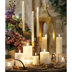 A variety of lit white candles of different heights are arranged on a table with glass votives, surrounded by a floral bouquet and a gold lantern in the background. The setting appears warm and elegant.