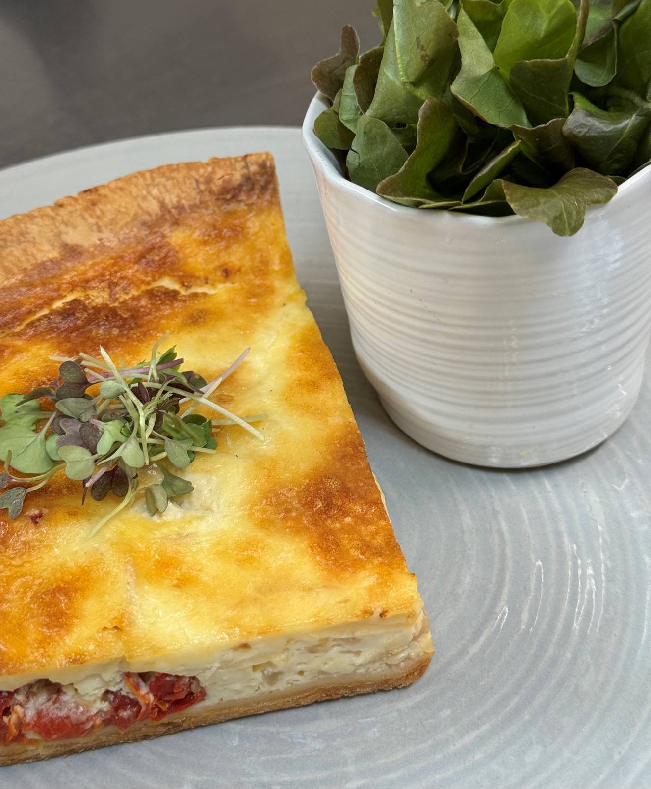 A slice of golden-brown quiche topped with microgreens is served on a gray plate next to a small white cup filled with fresh green salad leaves.