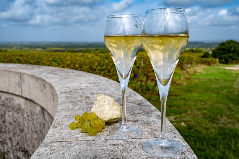 Two glasses of white wine, a small bunch of green grapes, and a stone rest on a curved stone ledge, with green vineyards and a blue sky in the background.