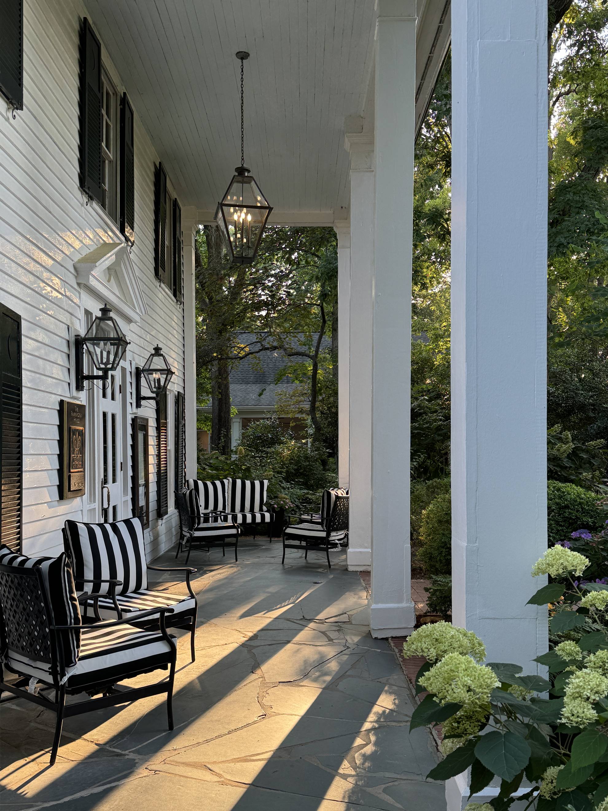 A sunlit porch with black-and-white striped chairs, a white wooden exterior, black shutters, hanging lanterns, and green hydrangeas in the foreground. Trees and a house are visible in the background.