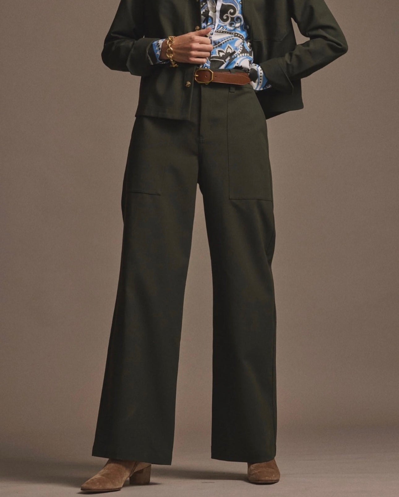 A person wearing dark wide-leg trousers, a brown belt, a patterned shirt, a dark blazer, and brown heeled shoes stands against a plain background. The face is not visible.