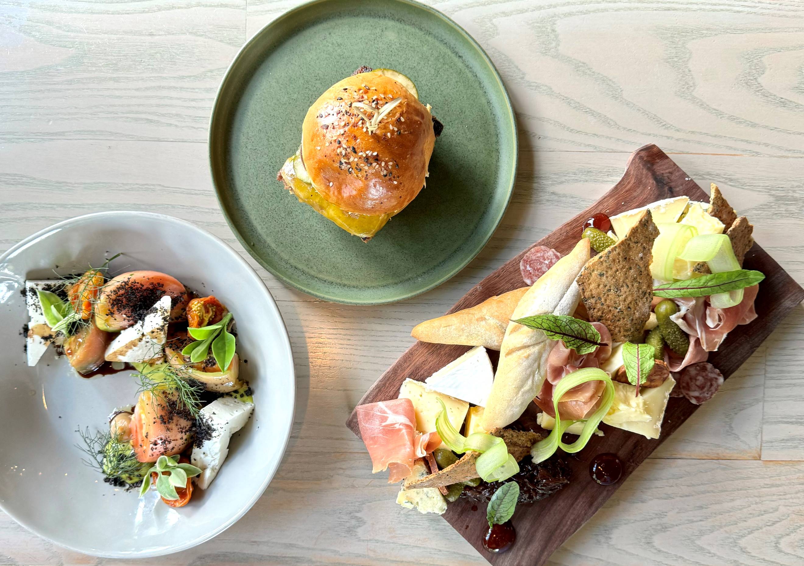 Three dishes on a light wooden table: a green plate with a sandwich, a white bowl with a colorful salad including tomatoes and cheese, and a wooden board with assorted cheeses, cured meats, crackers, and greens.