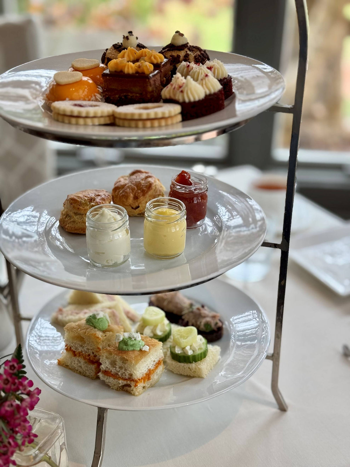 A three-tiered afternoon tea stand with assorted pastries and tarts on the top tier, scones with jam and cream in the middle, and finger sandwiches with cucumber and egg on the bottom tier, set on a white tablecloth.