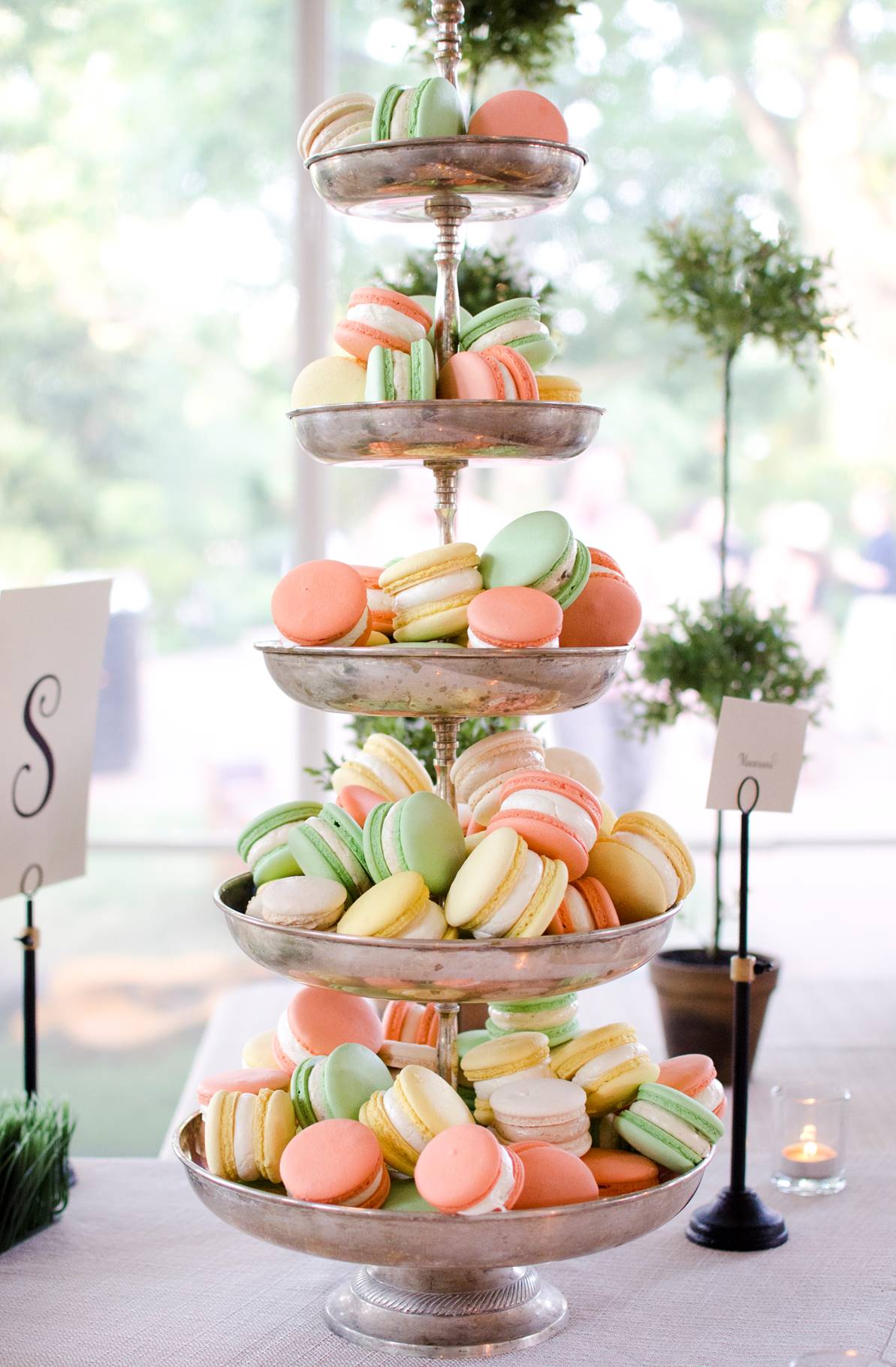 A four-tiered silver stand displays pastel-colored macarons in pink, green, yellow, and peach, arranged neatly on each level. The stand is set on a table with decorative greenery and cards nearby.