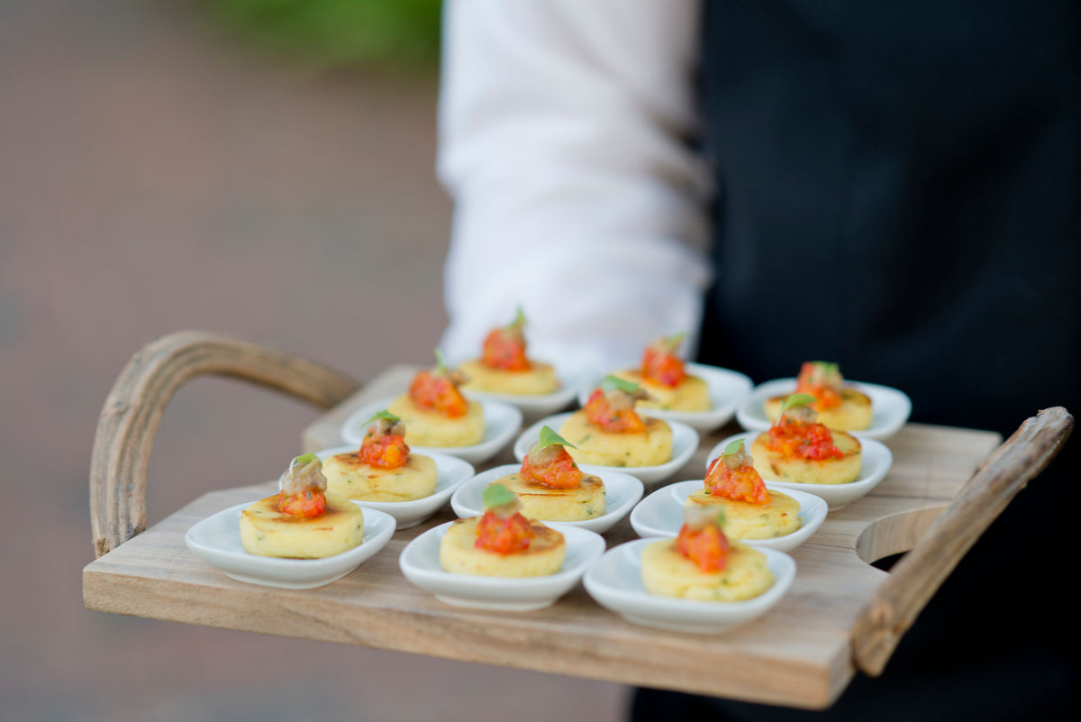 A person in formal attire holds a wooden tray with small white dishes, each containing an appetizer topped with a red and green garnish. The tray is presented outdoors.